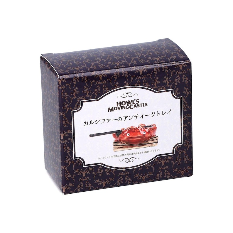 May include: A decorative box with a dark purple background and gold swirls. The box features a white label with the text "HOWL'S MOVING CASTLE" and Japanese text. Inside the label is a red tray with a black object.