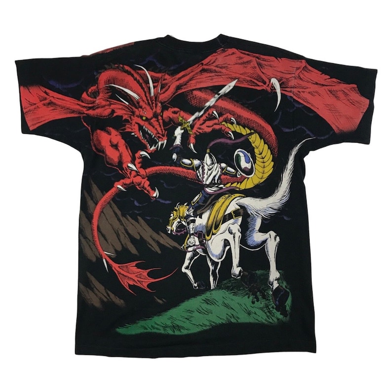 May include: Black t-shirt featuring a graphic print of a knight on a white horse battling a red dragon. The dragon has a long tail and sharp claws. The knight is wearing armour and wielding a sword. The background is a dark, stormy sky.