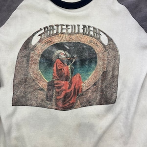 May include: A white t-shirt with a grey and black baseball style collar. The front of the shirt features a vintage graphic of a band called "Grateful Dead" with the words "A  Trips  For  Thursdays" printed above the graphic.