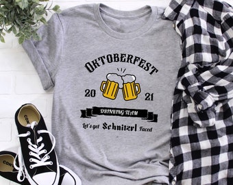 May include: Gray t-shirt with a black and white graphic design that says "Oktoberfest 20-21 Drinking Team Let's get Schnitzel Faced". The graphic includes two yellow beer mugs with foam on top.