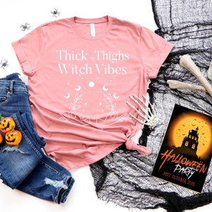 May include: A pink t-shirt with the text "Thick Thighs Witch Vibes" in white. The shirt features a graphic of two hands with moons, stars, and leaves.