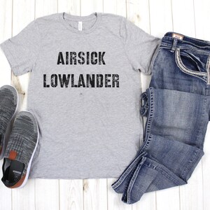 May include: A gray t-shirt with the text "AIR SICK LOWLANDER" printed in black. The shirt is laid flat on a wooden surface with a pair of gray sneakers and blue jeans.