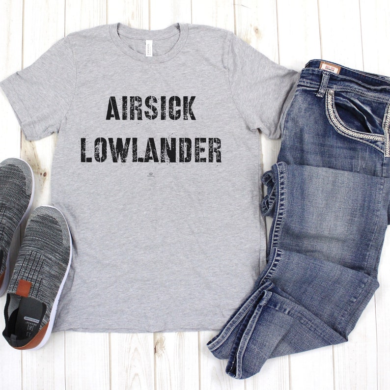 May include: A gray t-shirt with the text "AIR SICK LOWLANDER" printed in black. The shirt is laid flat on a wooden surface with a pair of gray sneakers and blue jeans.