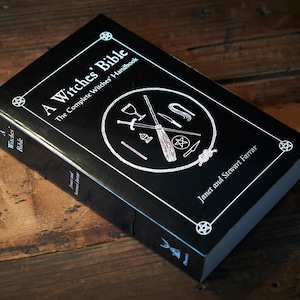 May include: A black book titled "A Witches' Bible: The Complete Witches' Handbook" by Janet and Stewart Farrar. The cover features a white pentacle and a circle with various witchy tools, including a broom, athame, chalice, and wand.