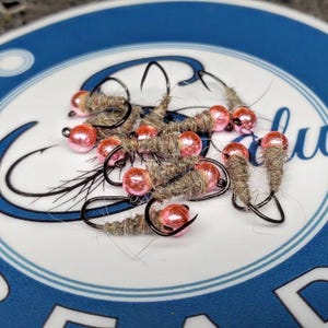 May include: A collection of fishing flies with gray bodies and pink bead heads. The flies are arranged on a white and blue patterned surface, showcasing their intricate design and craftsmanship. The hooks are black.