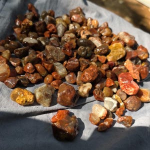 May include: A collection of small, polished, and raw river stones in various shades of brown, orange, red, and translucent white. The stones exhibit diverse textures, ranging from smooth and rounded to jagged and irregular, with some showing banding.