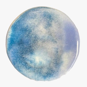 Puede incluir: Un posavasos circular con un acabado liso y brillante con un efecto de acuarela difuminado. La superficie muestra un degradado de azul profundo, azul cielo claro y tonos lavanda suaves, intercalados con sutiles reflejos blancos y una textura brillante.