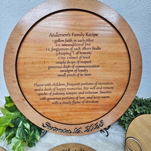 May include: A wooden round plaque with a laser engraved recipe for a family recipe called "Anderson's Family Recipe". The recipe is a list of ingredients for a happy marriage, including faith, love, honesty, trust, respect, communication, loyalty, and in-laws. The plaque is dated September 16, 2023, and includes the names "Cody & Michelle".