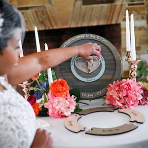 May include: A bride and groom participate in a wedding ceremony, placing a wooden heart puzzle piece into a circular wooden frame with the text "Our Path, Our Family, One Piece At A Time" and "Est. July 3, 2022".