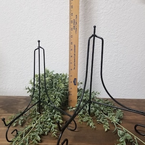 May include: Two black metal easel stands with a decorative scroll design on the base. The stands are on a wooden surface with green foliage.