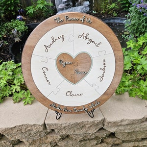 May include: A wooden round puzzle with a heart in the center. The puzzle pieces are labeled with names: Audrey, Abigail, Connor, Ryan, Sara, Jackson, and Claire. The text "The Pieces of Us" and "Make Our Family Whole" are written on the puzzle.