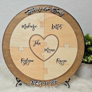 May include: A round wooden plaque with a heart-shaped puzzle piece in the center. The heart is divided into two pieces with the names "John" and "Megan" written on them. The plaque is surrounded by four puzzle pieces with the names "Makenzie", "Karter", "Rylynn", and "Rowyn" written on them. The plaque also has the text "Together We Make A Family" and "October 21, 2023" written on it.
