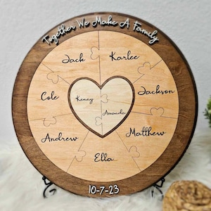 May include: A round wooden plaque with a heart-shaped puzzle in the center. The heart is made up of eight puzzle pieces, each with a name written on it: Jack, Karlee, Cole, Kenny, Amanda, Jackson, Matthew, and Ella. The plaque also has the text "Together We Make A Family" around the edge and the date "10-7-23" at the bottom.