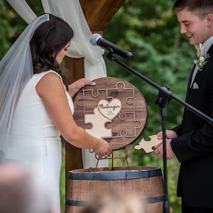 May include: A wooden heart-shaped puzzle with the text "Fleckinger Est. 8.25.23" on it. The puzzle is being put together by a bride and groom during their wedding ceremony.