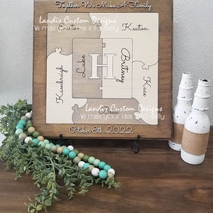 May include: A wooden puzzle with the names Kambreigh, Luke, Britney, and Kace, along with the letter H in the center. The puzzle is framed in a brown wood frame and has the text "Together We Make A Family" and "Landis Custom Designs" on it. The date "October 8th, 2022" is also on the puzzle.