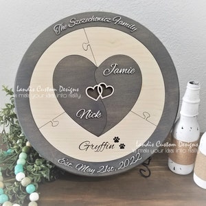 May include: A round wooden plaque with a gray stain and a light wood center. The plaque is divided into sections with a heart shape in the center. The heart is divided into two halves, one gray and one light wood. The names "Nick" and "Jamie" are written inside the heart. The words "The Szczechowicz Family" are written around the top edge of the plaque. The words "Landis Custom Designs" are written on the bottom left and right sides of the plaque. The words "Est. May 21st, 2022" are written on the bottom center of the plaque. The words "Gryffin" are written on the bottom right side of the plaque with paw prints.