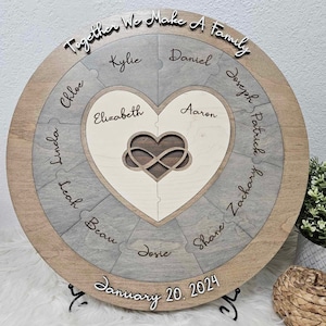 May include: A round wooden plaque with a heart-shaped puzzle in the center. The heart is made of two pieces, each with an infinity symbol carved into it. The plaque is surrounded by a ring of puzzle pieces, each with a name written on it. The names are: Linda, Chloe, Elizabeth, Kylie, Daniel, Joseph, Patrick, Zachary, Leah, Beau, Josie, Shane. The plaque also has the text "Together We Make A Family" and "January 20, 2024" written on it.