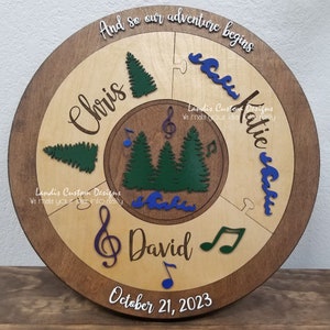 May include: A wooden round sign with a brown border and a light wood center. The sign is divided into four sections with a puzzle piece design. Each section has a different design: a tree, a wave, a musical note, and a name. The names are "Chris", "Katie", "David", and the date "October 21, 2023". The text "And so our adventure begins" is written at the top of the sign.