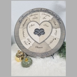 May include: A round wooden puzzle with a heart-shaped center and the text "God Builds A Family One Piece At A Time". The puzzle pieces are labeled with names, including "Marjorie & Ricky", "Christine & Irv", "Meaganne", "Alex", "Eric", "Andren", "Boots & Tiger", "Kimchi", "Zack", and "Thomas". The date "10/21/2022" is inscribed below the puzzle.