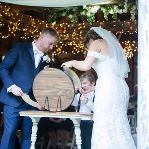 May include: A bride and groom sign a wooden guest book at their wedding ceremony. A young boy in a white shirt and bow tie stands next to them. The ceremony is taking place in a rustic barn with string lights hanging from the ceiling.