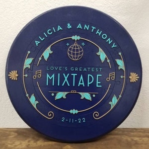 May include: A blue circular wooden sign with gold and teal accents. The sign reads "Alicia & Anthony Love's Greatest Mixtape 2-11-22" and features a gold disco ball graphic.