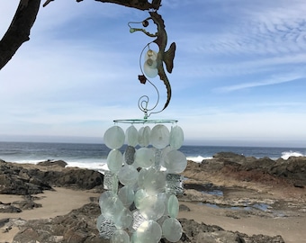 May include: A seahorse wind chime with a metal seahorse silhouette and a cascade of translucent, round, and square shell-like pieces. The wind chime hangs from a tree branch against a backdrop of the ocean and a blue sky.