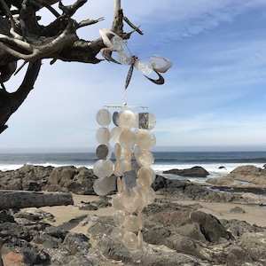 May include: A white and silver wind chime with a dragonfly design hanging from a driftwood branch. The wind chime is made of seashells and is hanging over a rocky beach.