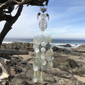 May include: A silver owl wind chime with a white and gray shell design hangs from a tree branch. The wind chime is made of metal and has a variety of different sized shells. The wind chime is hanging over a rocky beach.