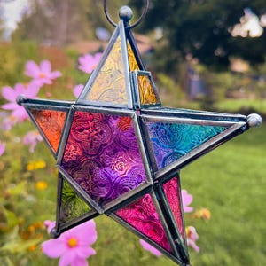 May include: A three-dimensional star-shaped hanging ornament constructed from multi-colored textured glass panels held together by dark metal soldering. The glass features vibrant shades of yellow, orange, purple, blue, and pink with embossed patterns.