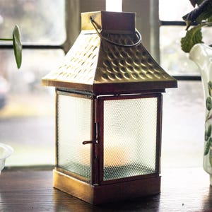 May include: A square metal lantern featuring a hammered gold-tone roof, a hinged door with a simple latch, and four clear glass panels with a textured grid pattern. A circular metal handle is attached to the top of the lantern frame.