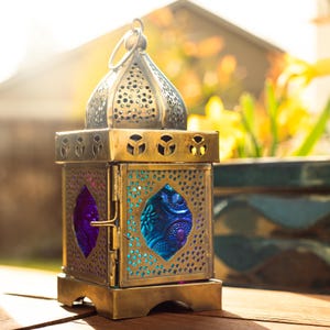 May include: A square metal lantern with a brass finish featuring a domed top with circular perforations. The sides have intricate cut-out patterns and inset panels of translucent blue and purple glass. A small metal ring sits at the very top.