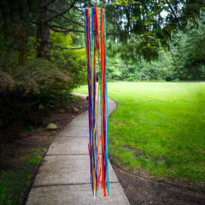 May include: A colorful windsock with multiple ribbons in shades of red, orange, yellow, green, blue, and purple hanging from a stick. The windsock is hanging in a grassy area with a paved walkway leading to it.