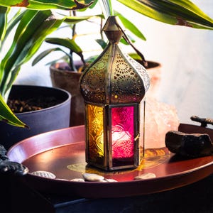 May include: A hexagonal metal lantern featuring an intricate filigree top and panels of textured yellow and magenta glass. The lantern has a pointed dome shape with a small metal ring at the apex for hanging, showcasing a metallic finish.