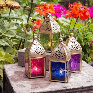 May include: Four decorative metal lanterns with intricate designs and colorful glass panels. The lanterns are arranged on a wooden surface with a green leafy background.