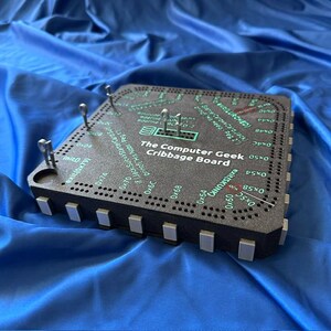 May include: A black cribbage board with the text "The Computer Geek Cribbage Board" in white. The board has a grid of holes and metal pegs. The board is on a blue surface.
