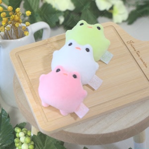 May include: Three plush frog toys in different colors: green, white, and pink. The frogs are sitting on a wooden cutting board with the text "Jaume Tomàs, Barcelona" engraved on it.