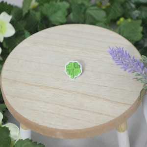 May include: A green four-leaf clover embroidered patch on a white background. The patch is sitting on a wooden surface.
