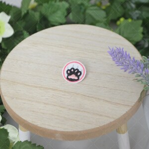 May include: A small, round, pink and white embroidered patch with a black paw print in the center. The patch is on a wooden surface.