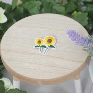 May include: Two embroidered sunflower patches on a light wood surface. The sunflowers are yellow with brown centers and green stems and leaves.