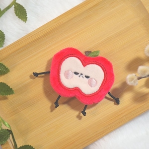 May include: A plush red apple toy with a white centre, embroidered with a grumpy face and small black legs and arms. The apple is on a wooden tray, with green leafy vines and fluffy white branches in the background.