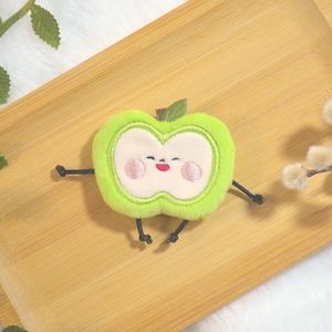 May include: A plush green apple character with a smiling face, arms, and legs, on a wooden tray. The apple has a light green outline, a white centre, and pink cheeks and mouth. A green leaf sits on top. Decorative branches with leaves and fluffy white flowers are in the background.