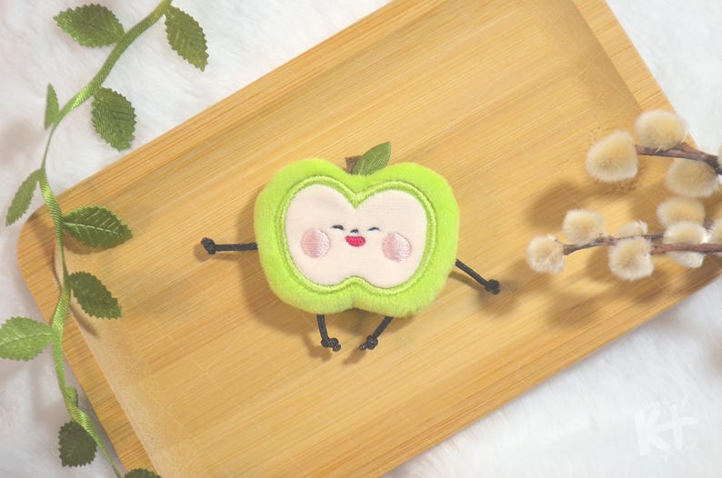 May include: A plush green apple character with a smiling face, arms, and legs, on a wooden tray. The apple has a light green outline, a white centre, and pink cheeks and mouth. A green leaf sits on top. Decorative branches with leaves and fluffy white flowers are in the background.