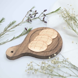 May include: Three golden brown bread rolls with a grid pattern on a small wooden chopping board with a handle. The board is surrounded by white and green flowers.