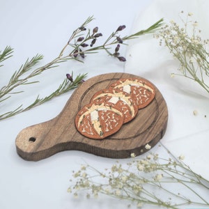 May include: A wooden serving board with three round, brown coasters resembling hot cross buns. The coasters have a white cross design and are surrounded by sprigs of rosemary and delicate white flowers.
