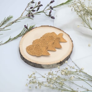 May include: Four golden fish-shaped cookies on a wooden slice, surrounded by white flowers and green sprigs.