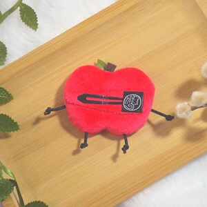 May include: A plush red apple-shaped toy with black arms and legs, and a black barrette. The apple has a small green stem and a black and white patch. The toy is on a wooden tray with decorative sprigs.