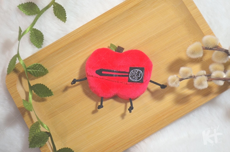 May include: A plush red apple-shaped toy with black arms and legs, and a black barrette. The apple has a small green stem and a black and white patch. The toy is on a wooden tray with decorative sprigs.