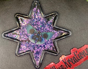 May include: A purple and blue glitter filled star-shaped resin ornament with a black butterfly inside. The ornament is on a black fabric background. The text "@KittyzKreations" is on a red glitter sticker in the lower right corner of the image.