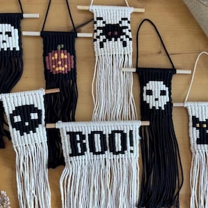 May include: A collection of seven small macrame wall hangings featuring Halloween-themed designs including a ghost, pumpkin, spider, skull, cat, and the text BOO!. Each piece is crafted from black or white cord on a wooden dowel with fringe.