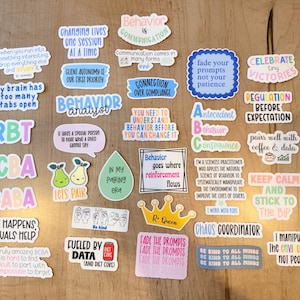 Puede incluir: Una colección de pegatinas coloridas con varias frases y gráficos relacionados con el análisis del comportamiento. Las pegatinas presentan textos como "Behavior Analyst", "Celebrate Tiny Victories" y "Fueled by Data". Las pegatinas están dispuestas sobre una superficie de madera marrón claro.